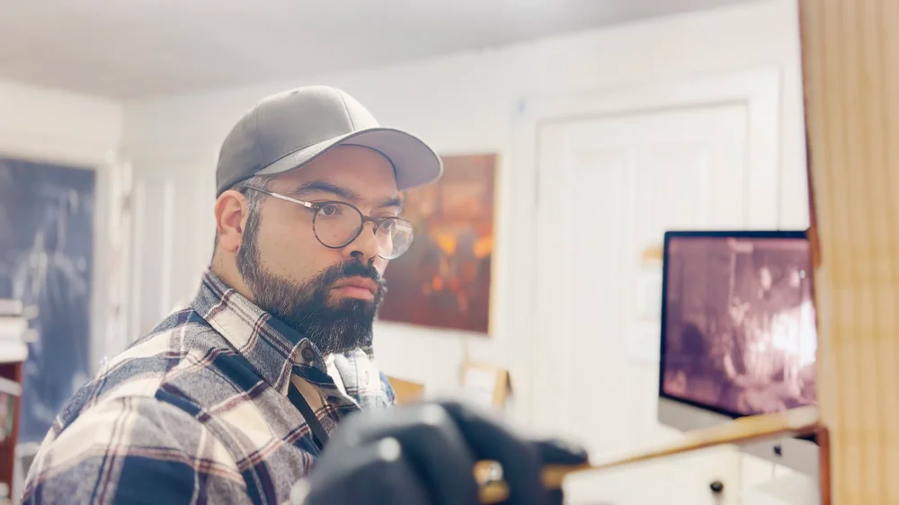 Raymond Bonilla in his studio, wearing glasses and a cap while focusing on a painting setup