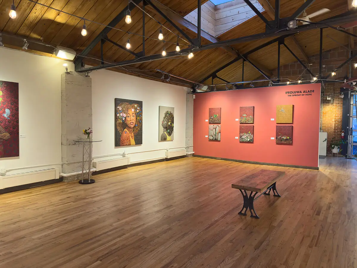 Corner installation view capturing Ifeoluwa Alade’s Oasis with its striking blue butterflies, Still I Rise (1) with yellow floral accents, and six nature-inspired square canvases including skulls, seedlings, and butterflies. A wooden bench sits in the center, grounded by soft natural lighting and exposed ceiling beams.