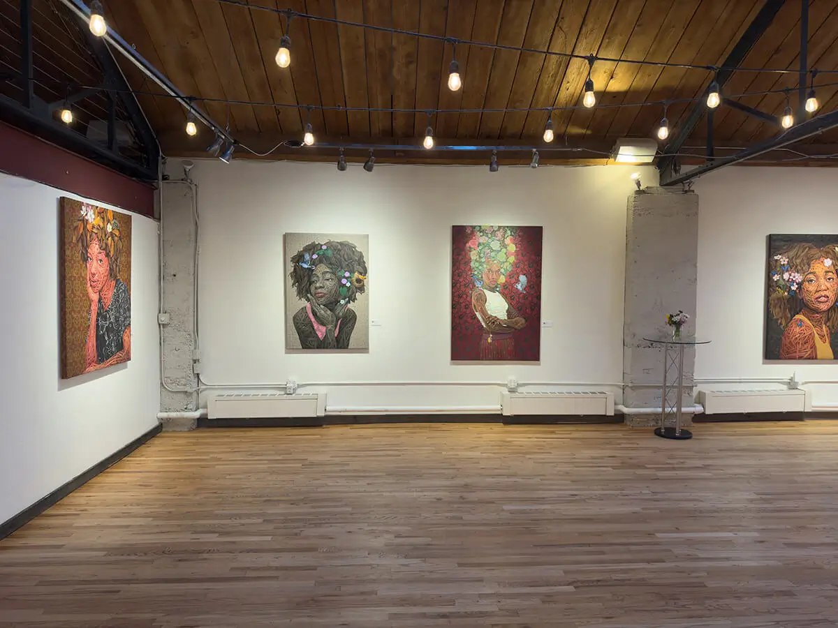 Installation photo from The Sprout of Hope, showing three large portraits by Ifeoluwa Alade. On the left, Beulah rests her chin on her hand, surrounded by blooming white flowers. Centered is Still I Rise (2) with the subject’s arms crossed, framed by pink and green blossoms. To the right is Still I Rise (1), a profile portrait with yellow accents. String lights and warm wood floors soften the space.