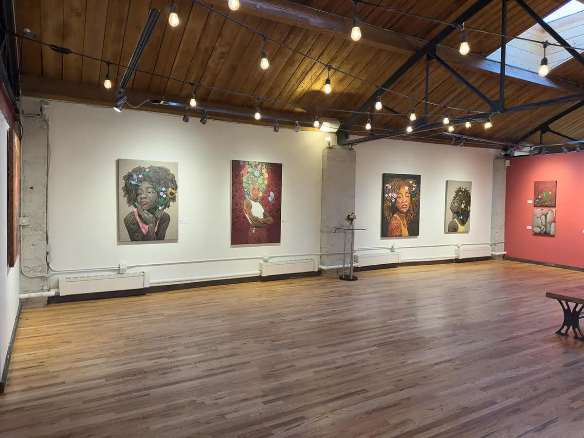 Gallery corner view showing four large portraits by Ifeoluwa Alade, including Beulah, Still I Rise (2) in red, Oasis with bright butterflies, and Still I Rise (1) in profile. To the right, the coral wall displays symbolic nature-based works including The Mustard Seed and Light in the Darkness I–IV. Skylight and string lighting bring softness and openness to the gallery setting.